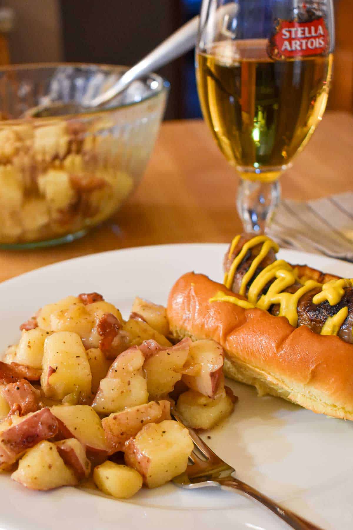A plate of German Potato Salad with a bratwurst on a bun, and a glass of beer in the background.