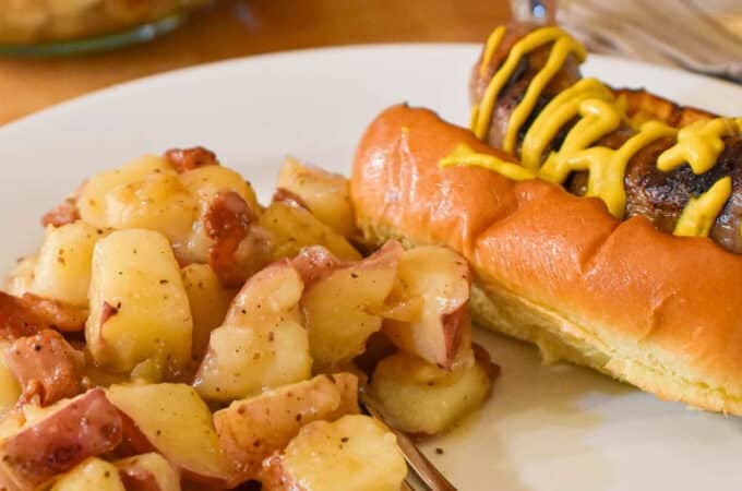 A plate of German Potato Salad with a bratwurst on a bun.